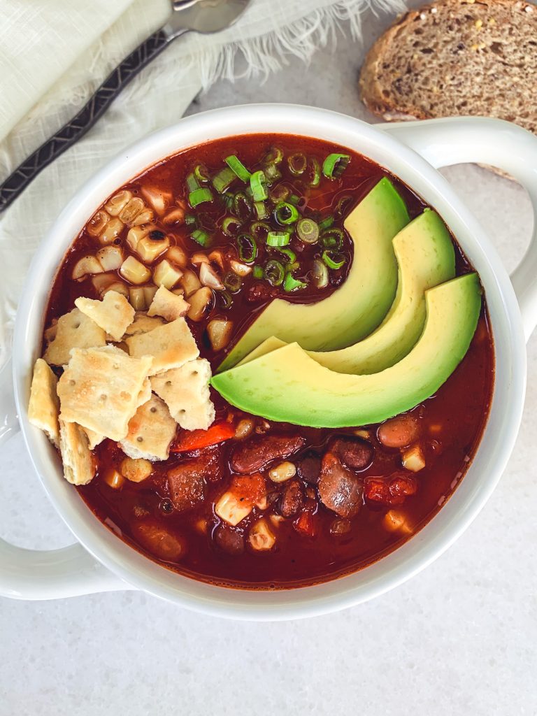 A large bowl of vegan 3 bean chili topped with avocado slices, roasted corn, green onions, and crushed up crackers.