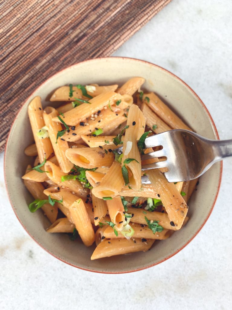 shot of a fork with penne noodles covered in a sesame tahini sauce with cilantro and scallions.