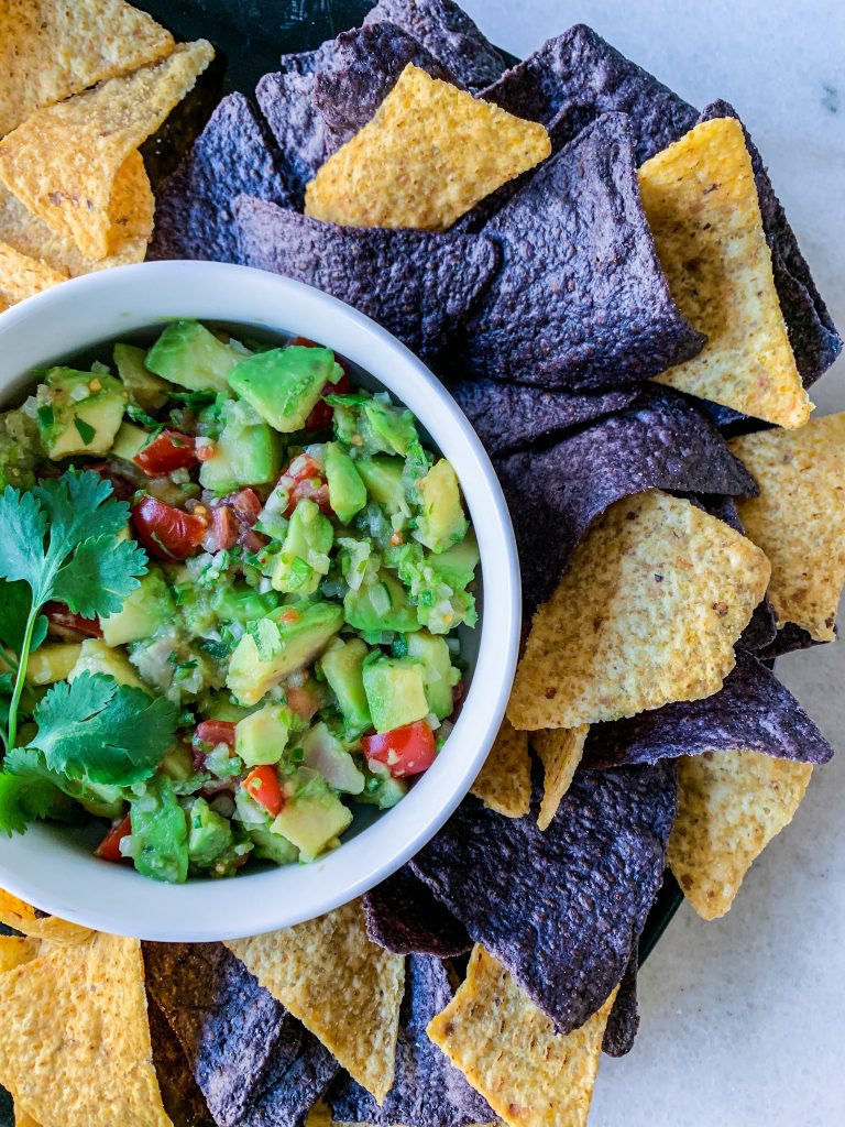 A bowl of fresh made guacamole with avocados, tomatoes, and cilantro; surrounded by yellow and blue corn tortilla chips.