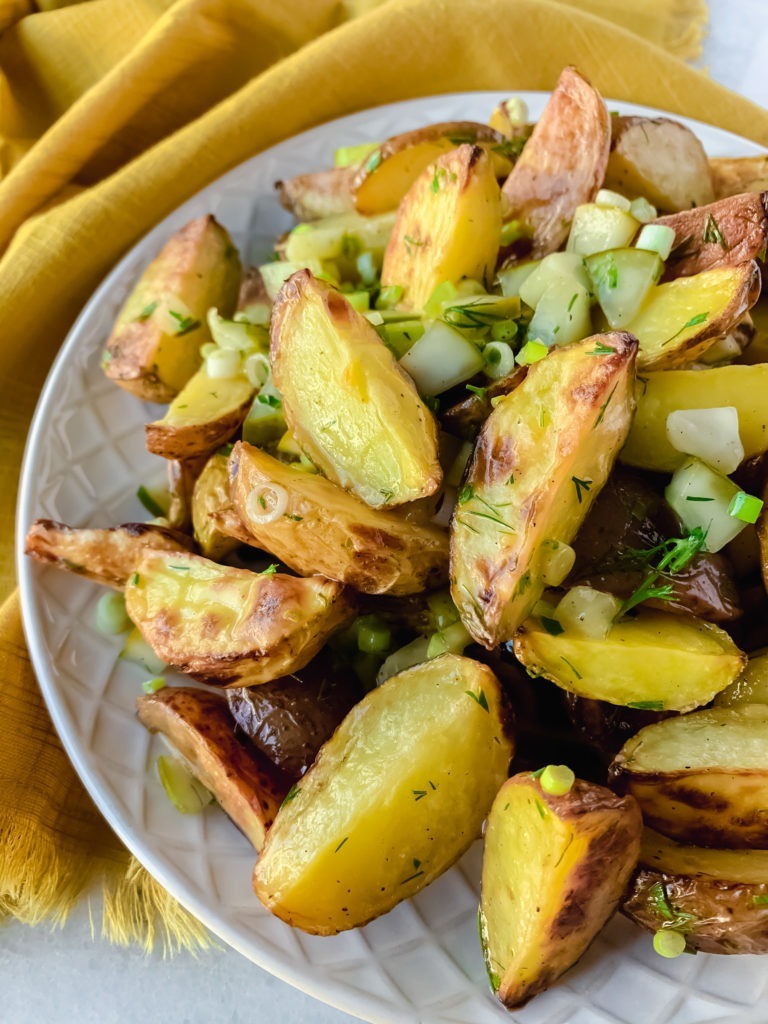 A plate full of crispy potato wedges coated in a maple mustard vinaigrette with fresh dill, green onions, and pickles.