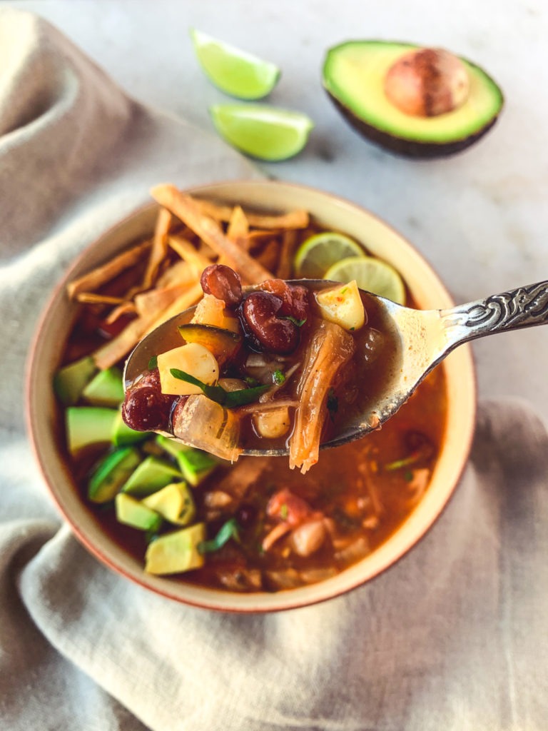 Spoon with jackfruit tortilla soup and garnished with tortilla strips and avocado.