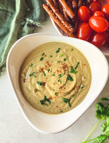 Bowl of creamy cilantro peanut hummus with pretzel sticks and cherry tomatoes for dipping.