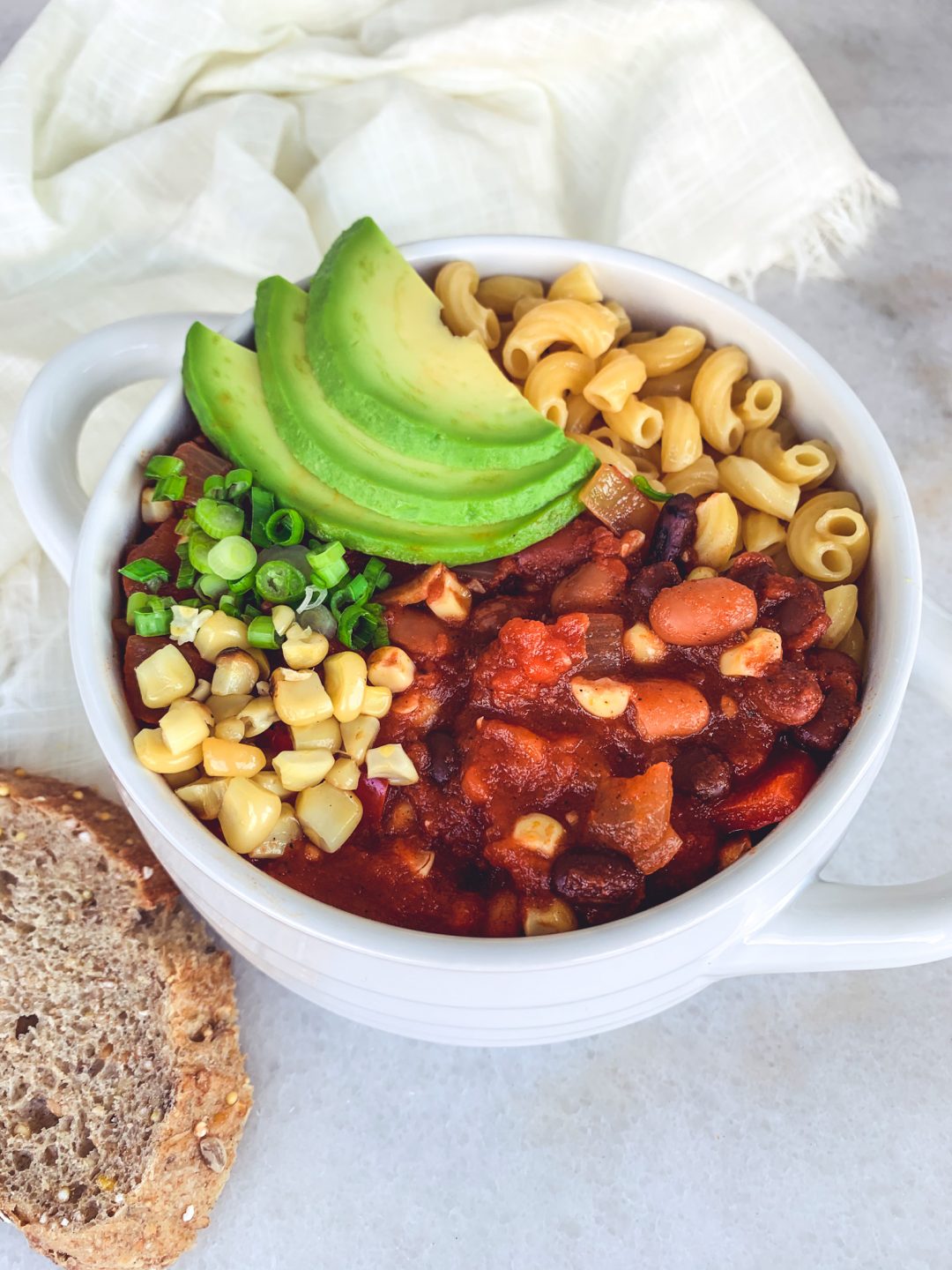 A bowl full of vegan chili made in a slow cooker using three different beans and veggies, and garnished with avocado, scallions, and roasted corn.
