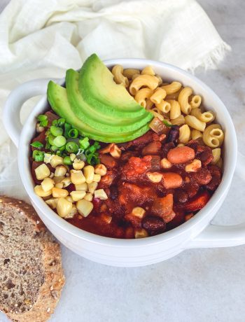 A bowl full of vegan chili made in a slow cooker using three different beans and veggies, and garnished with avocado, scallions, and roasted corn.